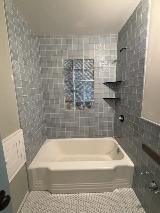 Blue-gray square tile tub surround centered on a glass block window, with two corner shelves