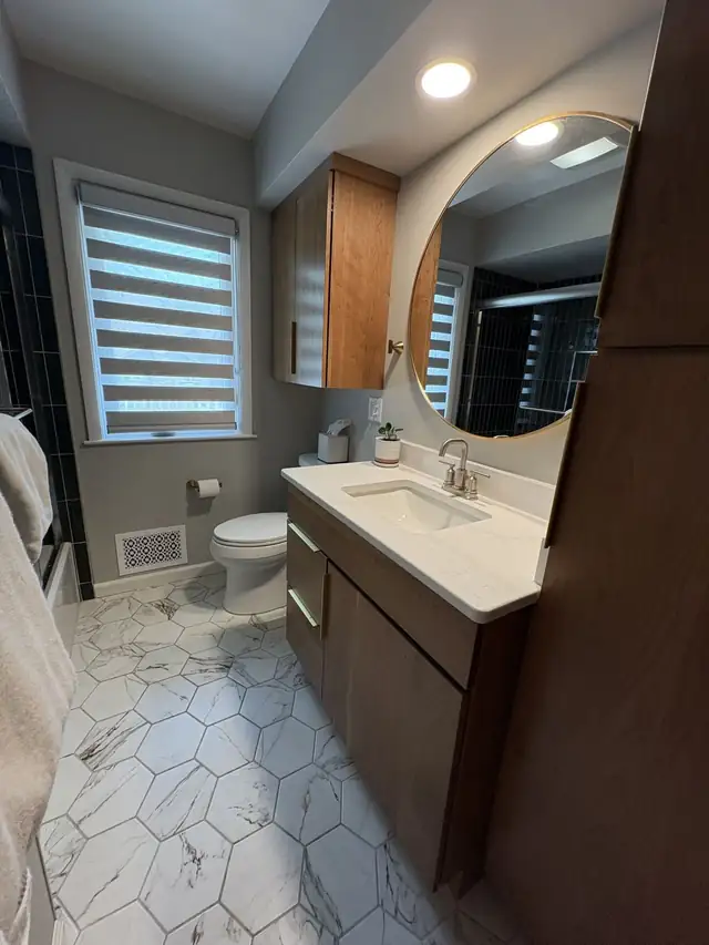 Remodeled bathroom with marble-look hexagon floor tile, a wood vanity with a white top and a round brass-framed mirror