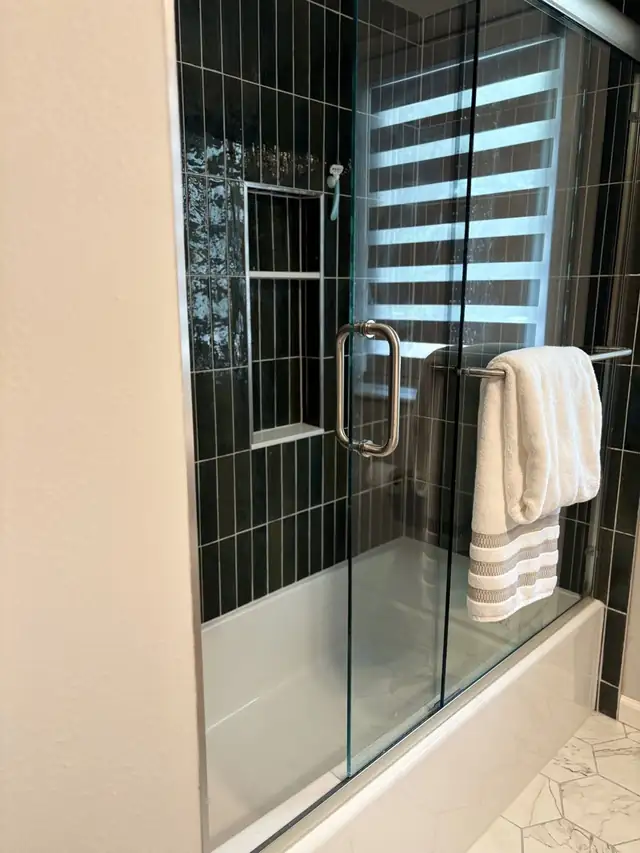Tub shower with dark green vertical tile and a glass door, with a white towel on the bar